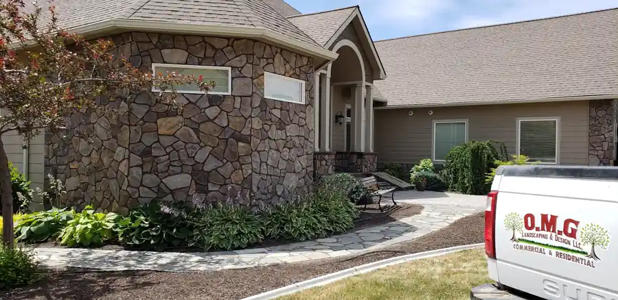 Stone-veneer house with a flagstone path and hosta bed, the OMG Landscaping truck door visible at the edge of the job