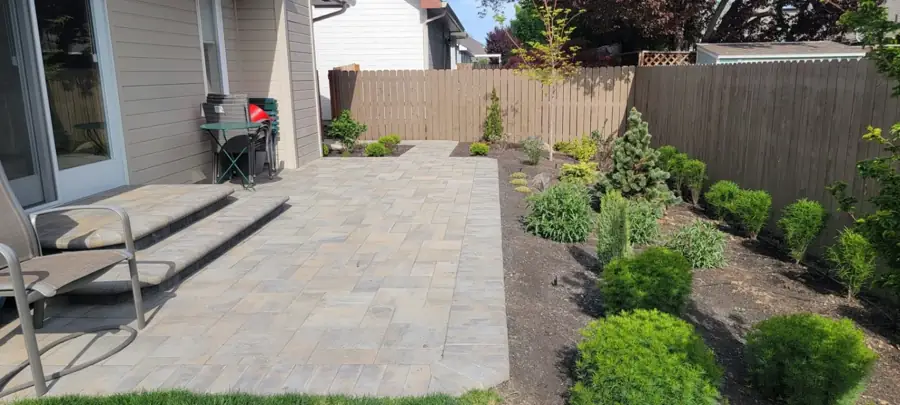 Backyard paver patio beside a shrub bed and cedar fence