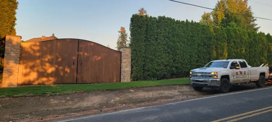 Arched wood driveway gate between stone pillars beside an arborvitae hedge, OMG pickup at the road